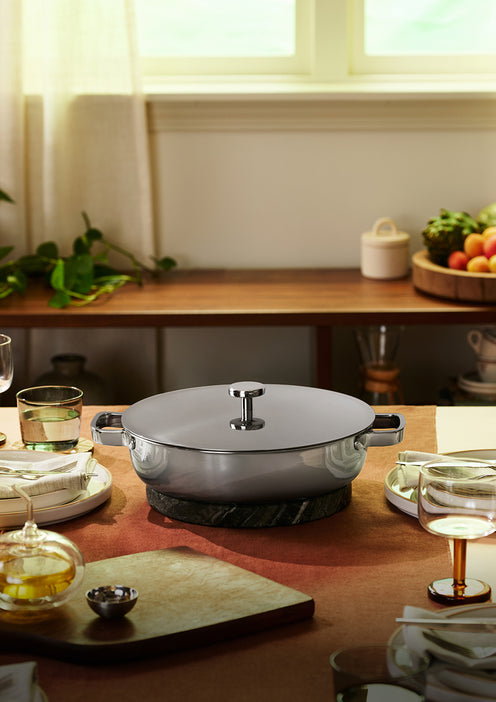 A covered stainless steel pot sits on a trivet at the center of a set dining table with plates, glasses, and food, in a sunlit room with a window and plants.