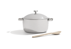 A stainless steel pot with a lid and side handles is placed next to a light-colored wooden spoon on a white background.