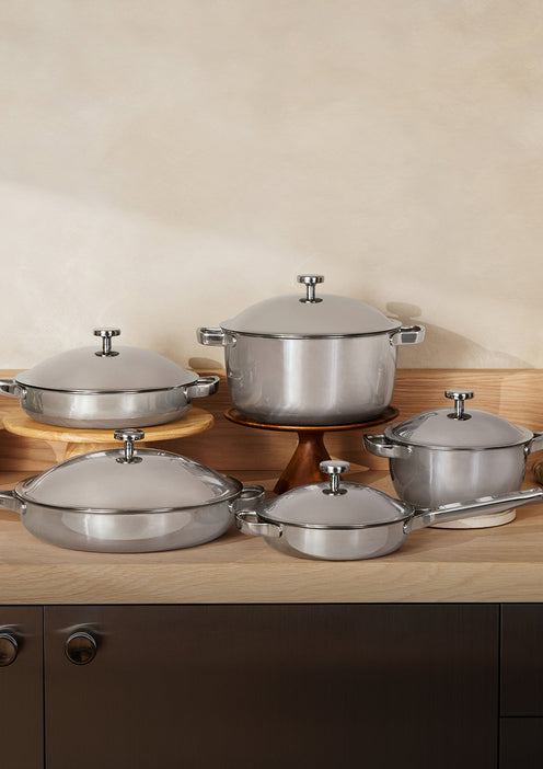 Five stainless steel pots and pans with lids are arranged on a wooden counter and shelf against a beige wall.