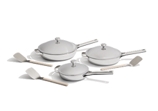 A set of three stainless steel pans with lids and four cooking utensils are arranged on a white background.
