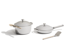 A set of stainless steel cookware including a lidded pot with a wooden spoon, a lidded frying pan, and a spatula, all on a white background.
