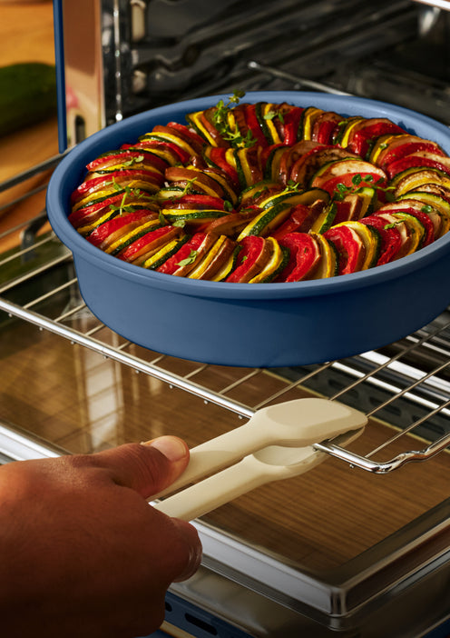 A hand uses tongs to slide a blue dish of sliced ratatouille onto an oven rack.