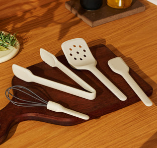A set of beige silicone kitchen tools, including a whisk, tongs, a slotted turner, and a spatula, arranged on a wooden cutting board.