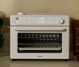 An off-white Our Place countertop oven with a digital display showing 230 degrees and 24:59, featuring function buttons for toast, air fry, reheat, roast, bake, broil, dehydrate, and proof.