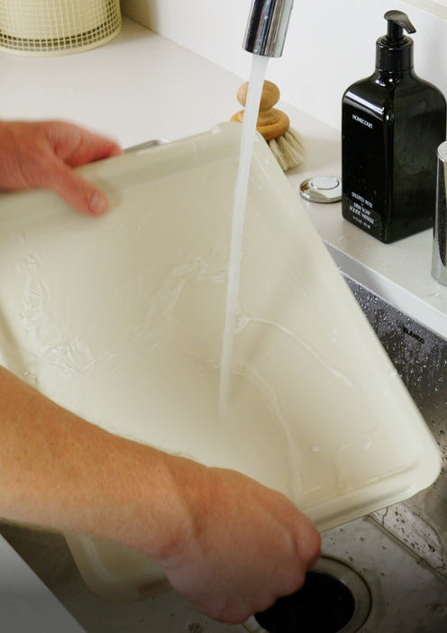 Hands washing a rectangular beige container under a kitchen faucet.