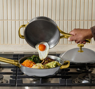 A hand is adding a fried egg from one pan into another pan filled with vegetables and noodles on a stove. A metal lid rests nearby.