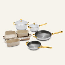 The Titanium Pro Cookware + Bakeware Set 2.0 features stainless steel cookware with gold-toned handles and lids, displayed with cream-colored ceramic bakeware on a checkered tray.