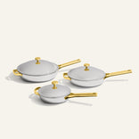 Three Titanium Pro Always Pan Trio 2.0 stainless steel frying pans of varying sizes, each with a matching lid and gold-toned handles and knobs.