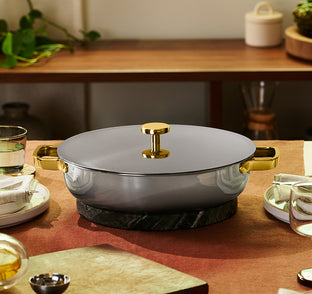A lidded gray and gold pot sits on a round black trivet on a set dining table with plates, glasses, and folded napkins.