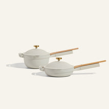 Two white cookware pieces with wooden handles and gold knobs, one deep pot and one shallow pan, are placed side by side on a light background.