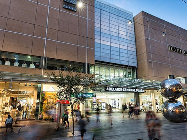 People walk past the entrance of Adelaide Central Plaza, with the David Jones store and two large reflective sculptures visible in front of the building.