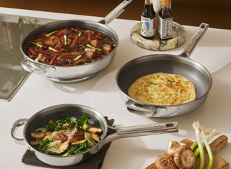 Three frying pans with cooked dishes on a kitchen counter, accompanied by condiments and ingredients.