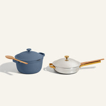 A blue Dutch oven with a wooden spoon rests beside a white sauté pan with a gold handle and lid, both on a light background.