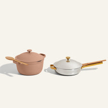 A pink dutch oven with a wooden spoon rests beside a white lidded skillet with a wooden handle and knob, set against a neutral background.