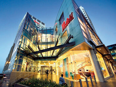 Modern glass-fronted shopping mall with a red Westfield sign, large entrance canopy, and multiple stories, shown at dusk with interior lights on.