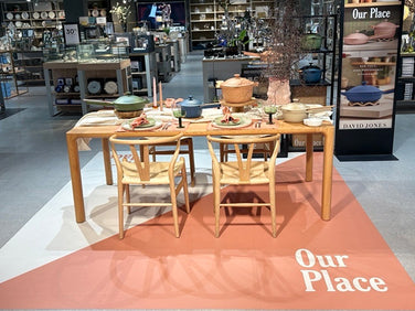Four wooden chairs and a dining table set with cookware and tableware are displayed in a store, with a sign reading Our Place on a mat and a standing banner promoting the brand.