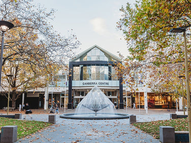 A glass-domed fountain sits in front of the Canberra Centre entrance, surrounded by trees with autumn leaves and modern lamp posts.