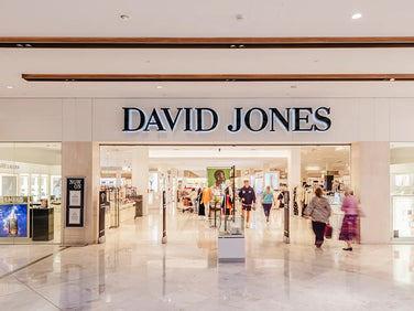Entrance to a David Jones department store in a shopping mall, with people walking in and out past displays and well-lit counters.