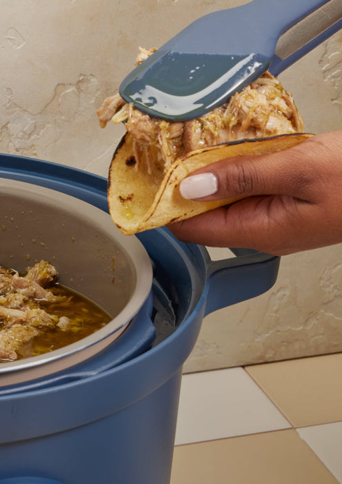 A hand holds a taco being filled with shredded meat using tongs, next to a blue slow cooker containing more meat and sauce.