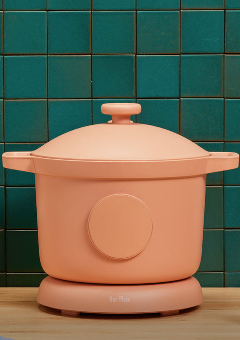 A peach-colored lidded pot sits on a matching base in front of a teal tiled wall, on a wooden countertop.