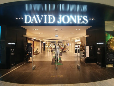 Entrance of a David Jones department store with illuminated signage, glass doors, mannequins, and displays visible inside.