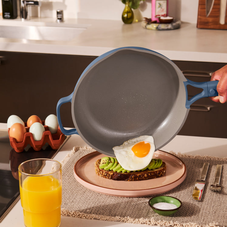 Person holding a frying pan over a plate with avocado toast and an egg, in a kitchen setting.