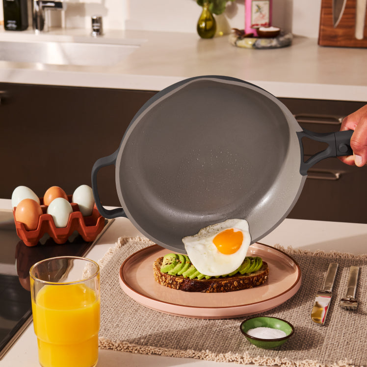 Person holding a frying pan with an egg on toast, next to a glass of orange juice in a kitchen.