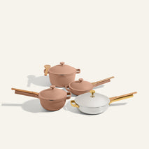 The Titanium & Ceramic Cookware Set, featuring a Perfect Pot with lid, pan with lid, sauté pan, and another pot in tan and white with gold handles, is displayed on a neutral background.