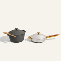 A black dutch oven with a wooden spoon and a white lidded frying pan with a wooden handle are placed side by side on a light background.