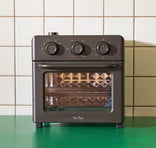 A compact, black countertop oven with three control knobs is placed on a green surface in front of a tiled wall.