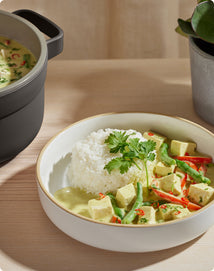 A bowl of white rice served with tofu, green beans, red chili, and green curry sauce, garnished with cilantro. A pot with more curry is nearby on a wooden table.