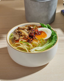 A bowl of ramen with noodles, bok choy, sliced mushrooms, half a soft-boiled egg, scallions, and chili oil, placed on a light wooden table.