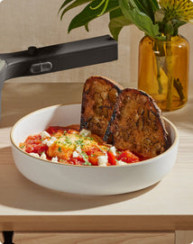 A bowl with two slices of toasted bread, tomato-based stew, and crumbled cheese sits on a light wooden surface near a vase with yellow flowers.