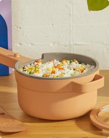 A beige saucepan filled with cooked rice mixed with corn, peas, and carrots sits on a wooden surface next to a lid and a wooden spoon.