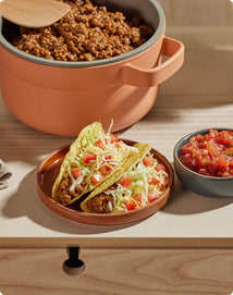Two hard shell tacos with ground meat, shredded lettuce, tomato, and cheese on a plate, with a bowl of salsa and a pot of taco filling in the background.