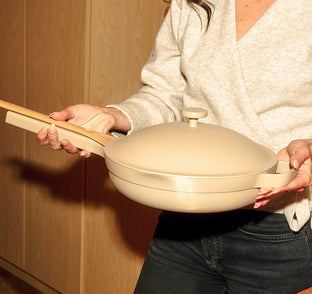 A person wearing a light sweater holds a beige frying pan with a lid and wooden handle in a kitchen setting.