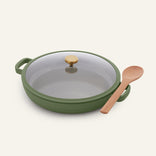 The Dual Handle Always Pan, a versatile green nonstick pan featuring a glass lid and elegant gold handle, sits next to a wooden spoon on a light background. This non-toxic cookware blends style and functionality to meet all your kitchen needs.