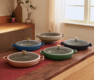 Five shallow, round, lidded pots in different colors are displayed on a kitchen island with a red runner and wooden cabinets in the background.
