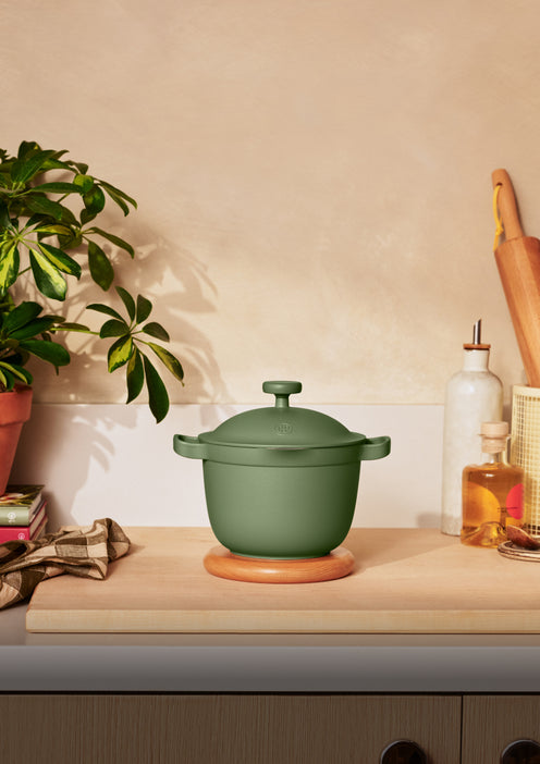 A green lidded pot sits on a wooden trivet on a kitchen counter, surrounded by a plant, kitchen towel, bottles, and utensils.