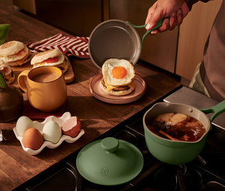 A person places a fried egg on an English muffin beside a mug, eggs, and a saucepan with liquid and food simmering on a stovetop.
