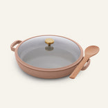The Dual Handle Always Pan, featuring a beige nonstick finish, glass lid with a gold knob, and paired with a wooden spoon resting on its handle, is pictured on a light background—making it an ideal choice for everyday cooking.