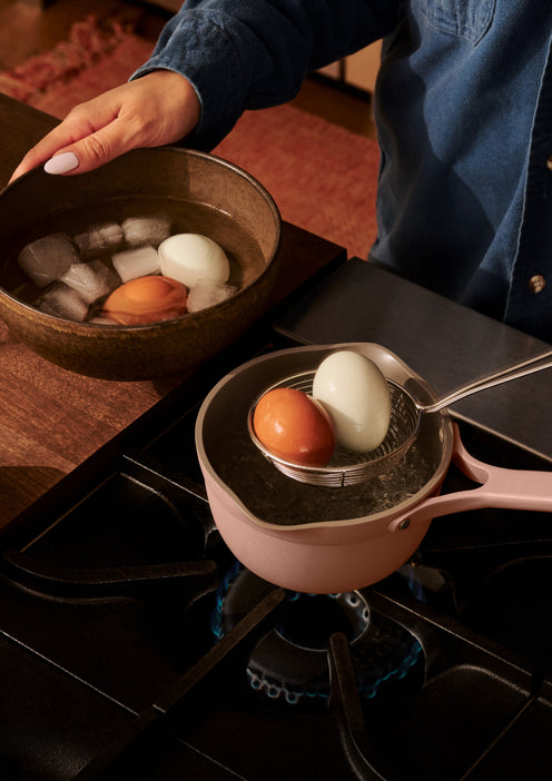 A person transfers boiled eggs from a pot of hot water on a stove into a bowl of ice water using a spoon.