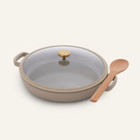 Versatile and non-toxic, the beige Dual Handle Always Pan features handles and a glass lid with a gold knob, and comes with a wooden spoon placed on the right side.