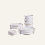 The Complete Plates & Bowls Set features white, chip-resistant ceramic dinnerware with gold rims—including plates, bowls, and small dishes—arranged in neat stacks on a neutral background.