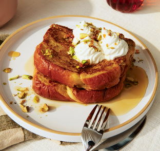 Two slices of French toast stacked on a plate, topped with whipped cream, chopped nuts, and syrup, with a fork and knife beside the plate.
