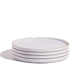 A stack of four white ceramic plates with thin rims, placed on a white surface against a plain background.