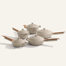 The Cookware Set+ features five beige non-stick pots and pans with matching lids and light brown handles, neatly arranged on a plain light background.