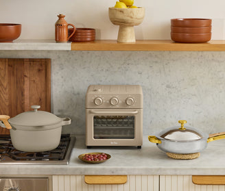 A beige air fryer oven sits on a marble countertop between a covered pot on a stove, a bowl of grapes, and a lidded pan; shelves with bowls and fruit are above.