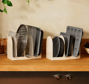 Two off-white kitchen organizers holding various metal baking pans, cooling racks, and muffin tins on a wooden countertop.