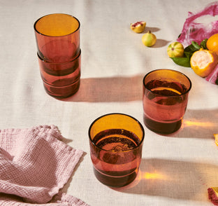 Stacked amber-colored glass tumblers on a light surface with fruits and a pink cloth.
